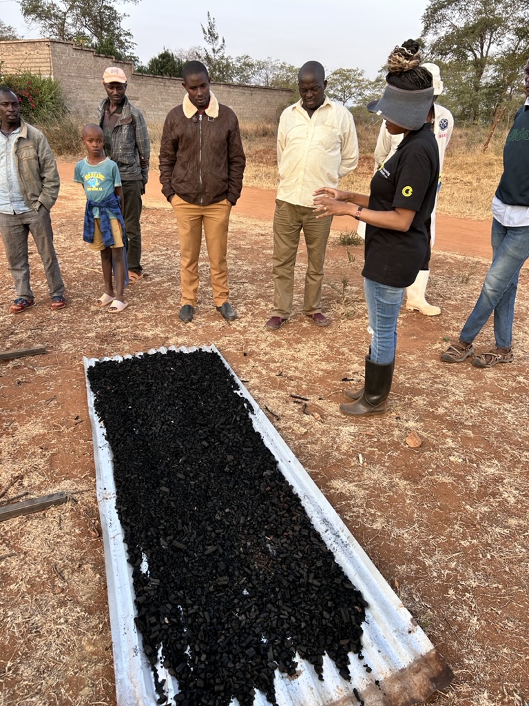 A Consuming Carbon team member explaining the properties and benefits of biochar to a group of local farmers gathered around a sample tray.