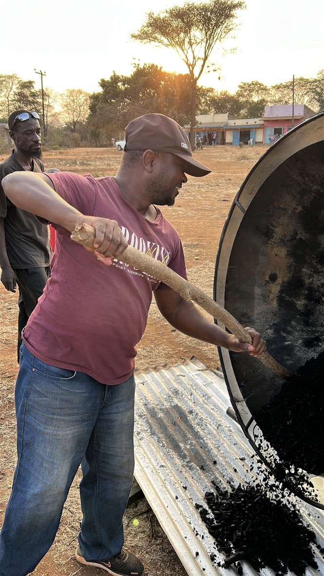 A team member shovelling freshly quenched biochar from the kiln onto corrugated metal sheeting at dusk, with a golden sky behind.