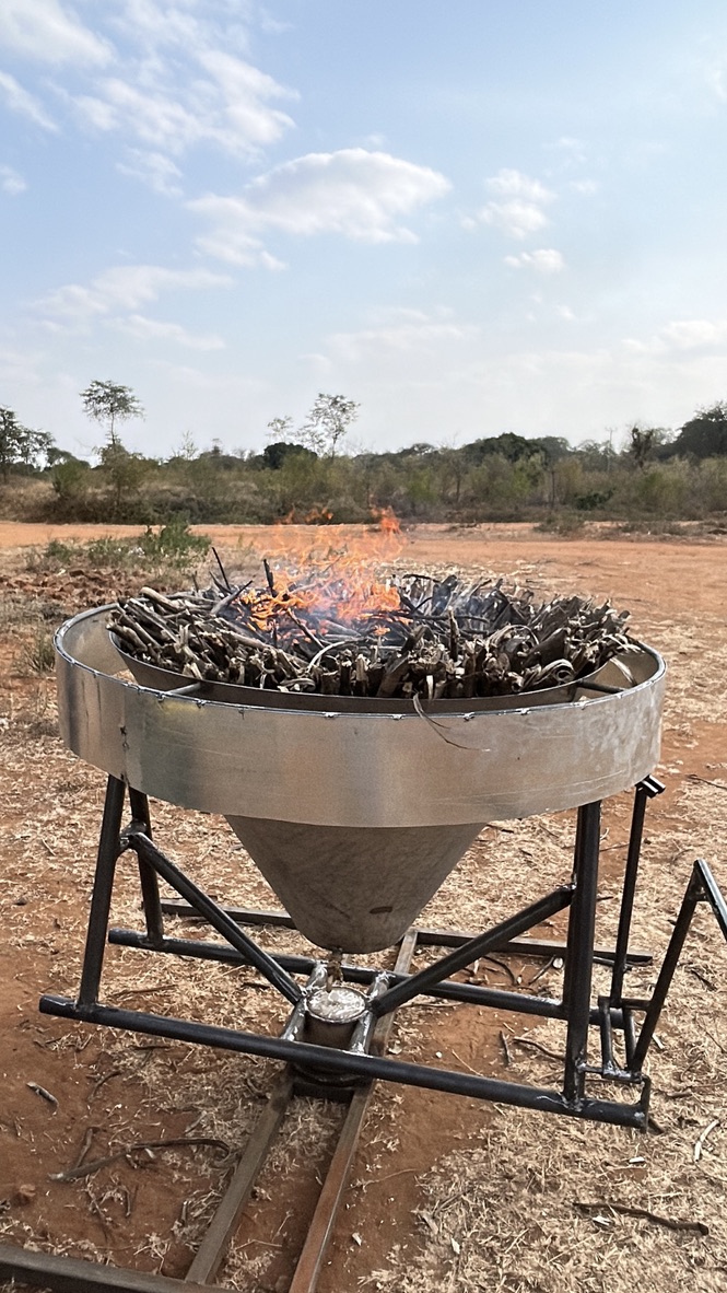 A Kontiki-style biochar kiln with active flames burning woody biomass feedstock on red laterite soil at Wamunyu.